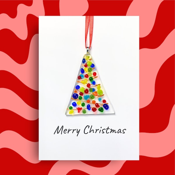 Puede incluir: Un adorno de cristal triangular con cuentas incrustadas de colores rojo, azul, verde y amarillo, suspendido por una cinta roja. El adorno se muestra sobre una tarjeta blanca con el texto Merry Christmas impreso en negro.