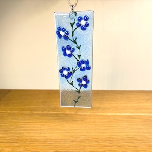 May include: A rectangular fused glass pendant featuring a light blue background with a subtle shimmer. A dark green vine pattern climbs vertically, adorned with five-petaled blue flowers that have white centres. A small metal loop is at the top.