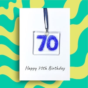 May include: A square glass ornament featuring the number 70 in textured blue glitter, suspended by a dark blue ribbon. The ornament is attached to a white card with the printed text Happy 70th Birthday centred at the bottom of the card.