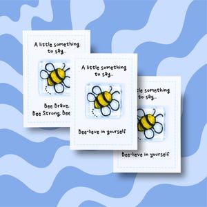 May include: Three white cards featuring a cartoon bee design. Each card has the text "A little something to say..." and "Bee-lieve in yourself" or "Bee Brave, Bee Strong, Bee". The background is a light blue wavy pattern.