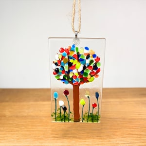 May include: A rectangular fused glass hanging ornament featuring a brown tree trunk with a canopy of multicoloured glass pieces in red, blue, green, yellow, and white. The base shows small colourful glass flowers on thin stems and green dots.