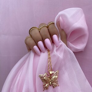 May include: A gold butterfly pendant necklace with a delicate, intricate design. The necklace is hanging from a hand with pink nail polish.
