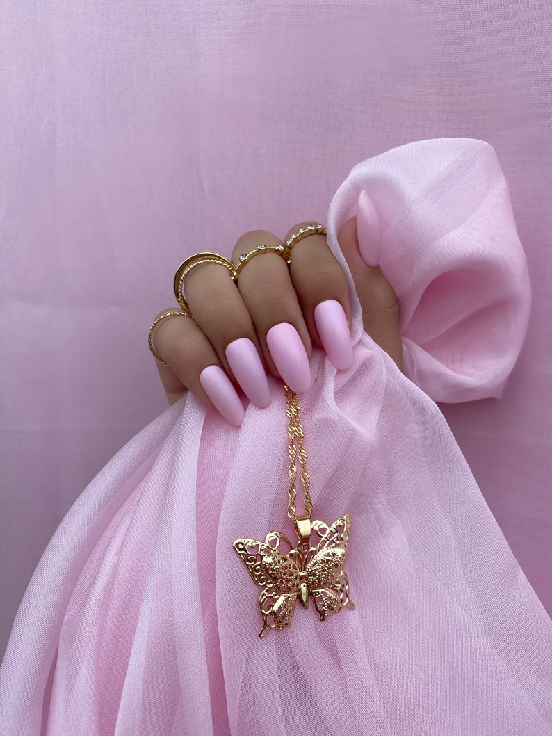 May include: A gold butterfly pendant necklace with a delicate, intricate design. The necklace is hanging from a hand with pink nail polish.