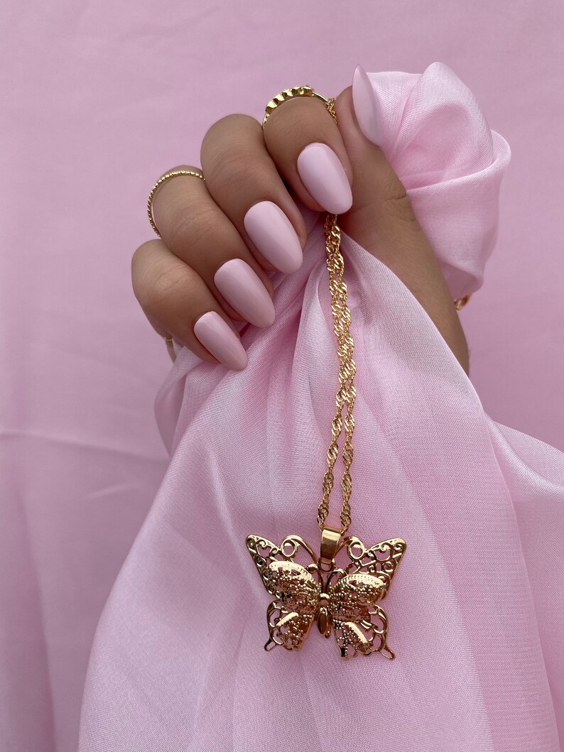 May include: A gold chain necklace with a delicate butterfly pendant. The necklace is draped over a hand with pink nail polish.
