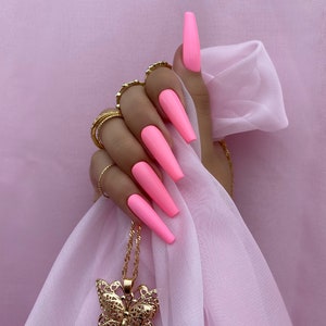 May include: A hand with long, bright pink nails is holding a gold butterfly pendant necklace. The necklace is hanging from the hand and the butterfly is in the center of the image.