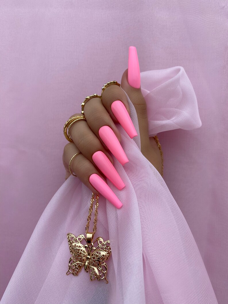 May include: A hand with long, bright pink nails is holding a gold butterfly pendant necklace. The necklace is hanging from the hand and the butterfly is in the center of the image.