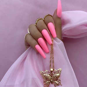 May include: A gold chain necklace with a large, ornate butterfly pendant. The necklace is hanging from a hand with long, pink acrylic nails.