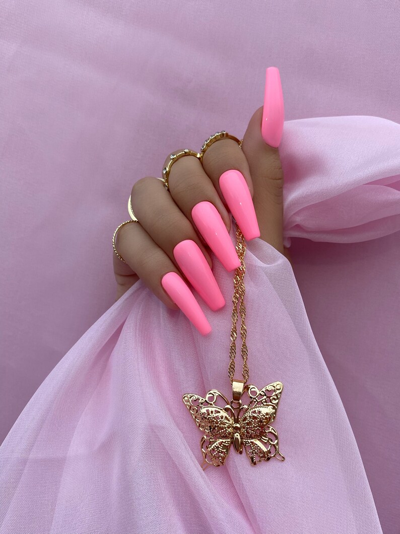 May include: A gold chain necklace with a large, ornate butterfly pendant. The necklace is hanging from a hand with long, pink acrylic nails.