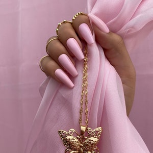 May include: A gold butterfly pendant necklace with a delicate chain. The necklace is hanging from a hand with pink long nails.