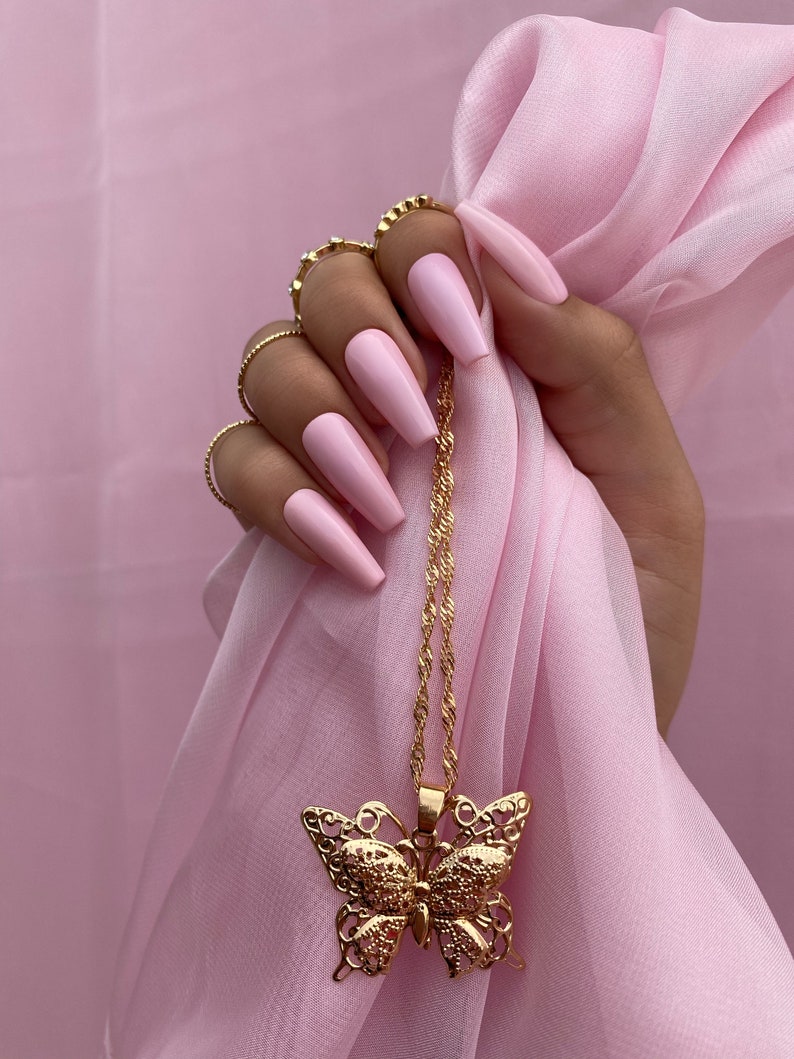May include: A gold butterfly pendant necklace with a delicate chain. The necklace is hanging from a hand with pink long nails.