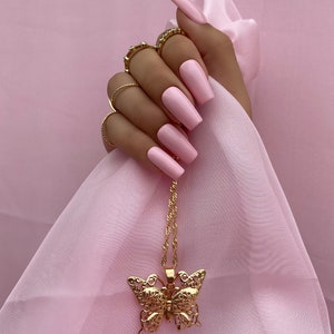 May include: A gold butterfly pendant necklace with a delicate chain. The necklace is hanging on a pink fabric background.