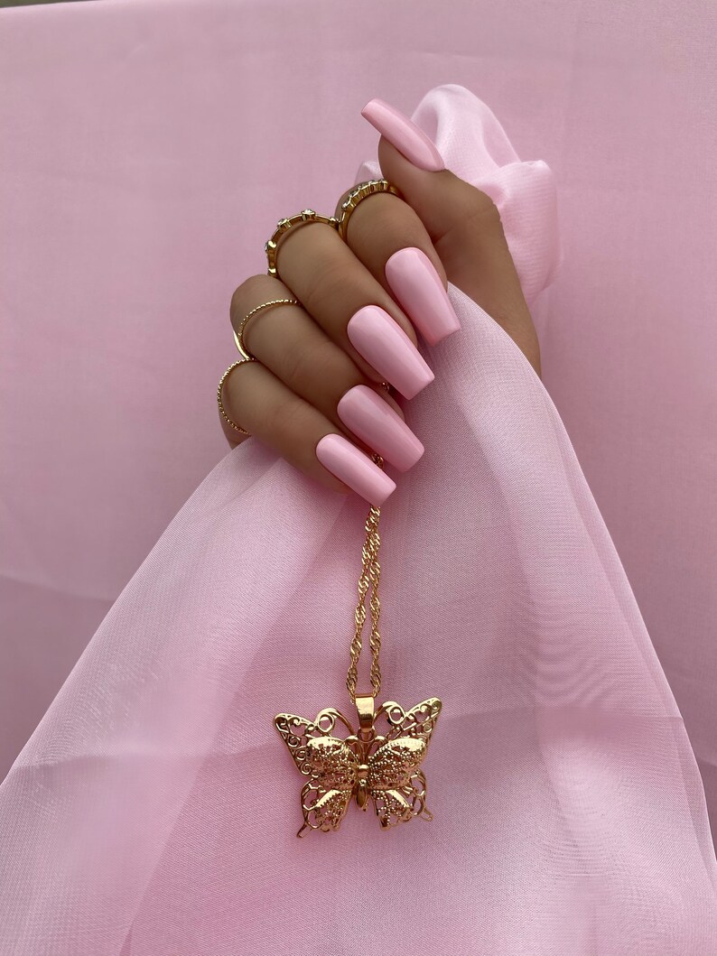 May include: A gold butterfly pendant necklace with a delicate chain. The necklace is hanging on a pink fabric background.