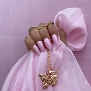 May include: A gold butterfly pendant necklace with a delicate filigree design. The necklace is hanging on a pink fabric background.