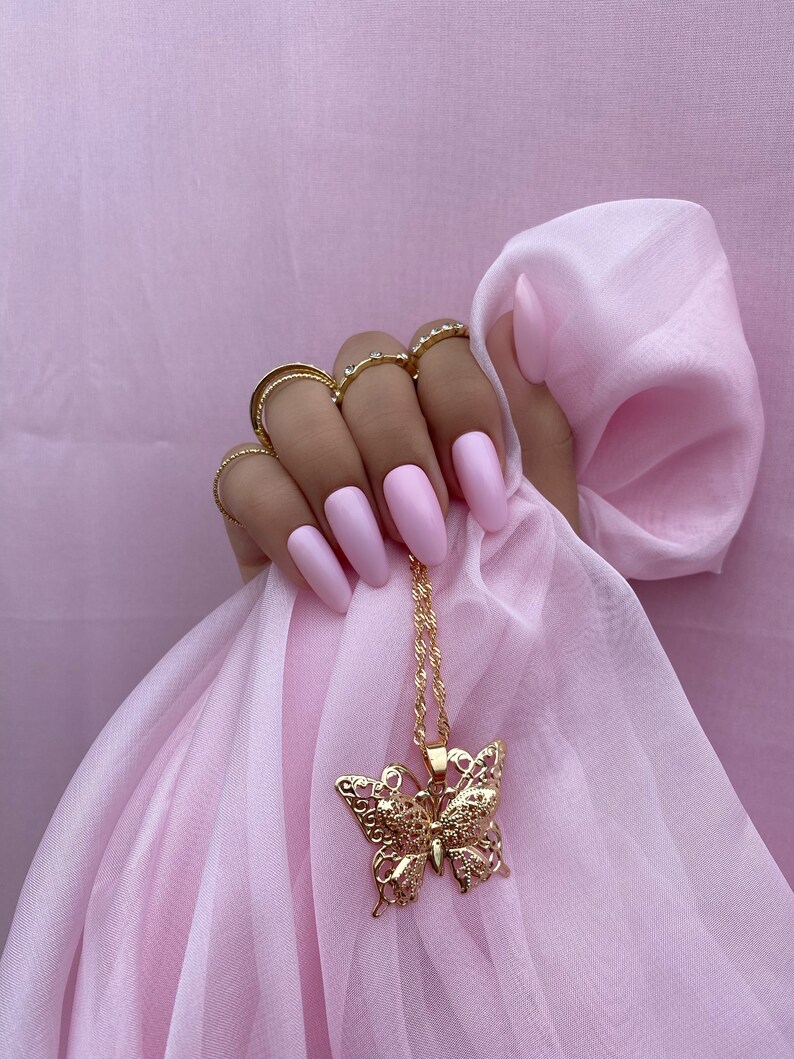 May include: A gold butterfly pendant necklace with a delicate filigree design. The necklace is hanging on a pink fabric background.