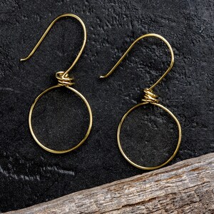 May include: A pair of gold-coloured hoop earrings. Each earring has a circular hoop design with a wrapped wire detail at the top where the hook is attached. The earrings are displayed on a dark, textured surface.