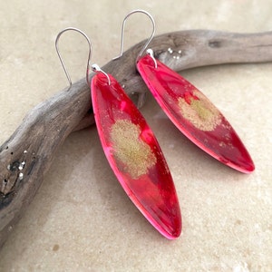 May include: A pair of long, oval-shaped earrings with a red and gold resin design. The earrings have silver hooks.