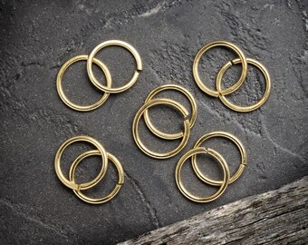 May include: A collection of eight gold-colored metal jump rings of various sizes. The rings are arranged on a dark gray textured surface.