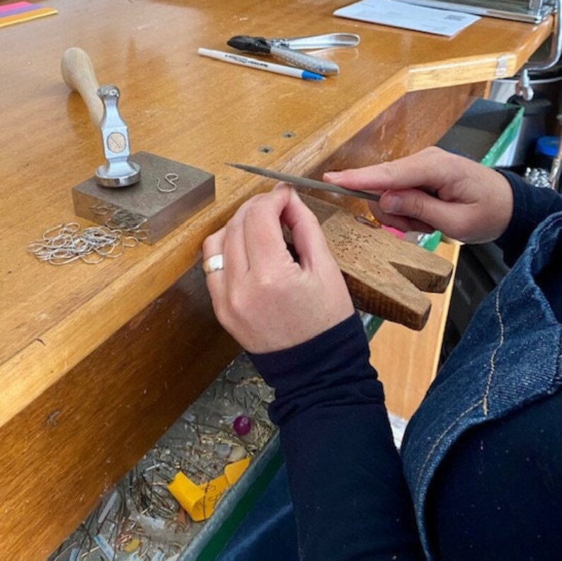 May include: A person working on a piece of jewelry with a file on a wooden workbench. There are tools and supplies on the workbench, including a hammer, a small block of wood, and a pile of metal rings.