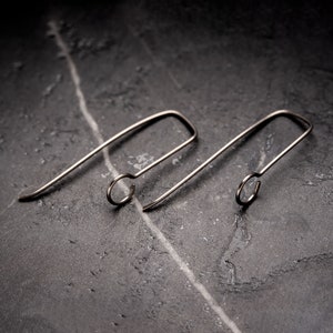May include: Two silver-coloured earrings with a minimalist design. Each earring has a curved, elongated shape with a small circular loop at the end. They are presented on a dark, textured surface, highlighting their sleek, modern style.