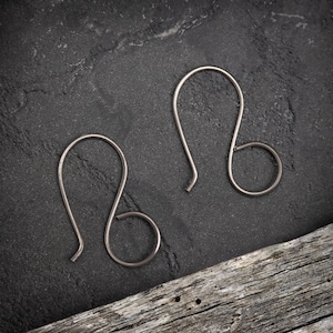 May include: Two silver-coloured, minimalist earring hooks. Each hook has a curved top, a looped middle, and a straight bottom. The earrings are displayed on a dark grey, textured surface with a piece of driftwood.