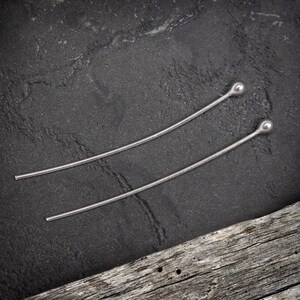 May include: Two silver hair pins with rounded ends. The pins are lying on a dark grey textured surface.