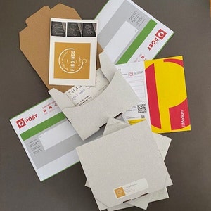 May include: A flat cardboard box with a white label that says "Thank you" and "Please leave your printed address details below". The box is surrounded by various packaging materials, including a brown envelope, a green and white envelope with the word "POST" on it, and a yellow and red envelope with the word "POST" on it.