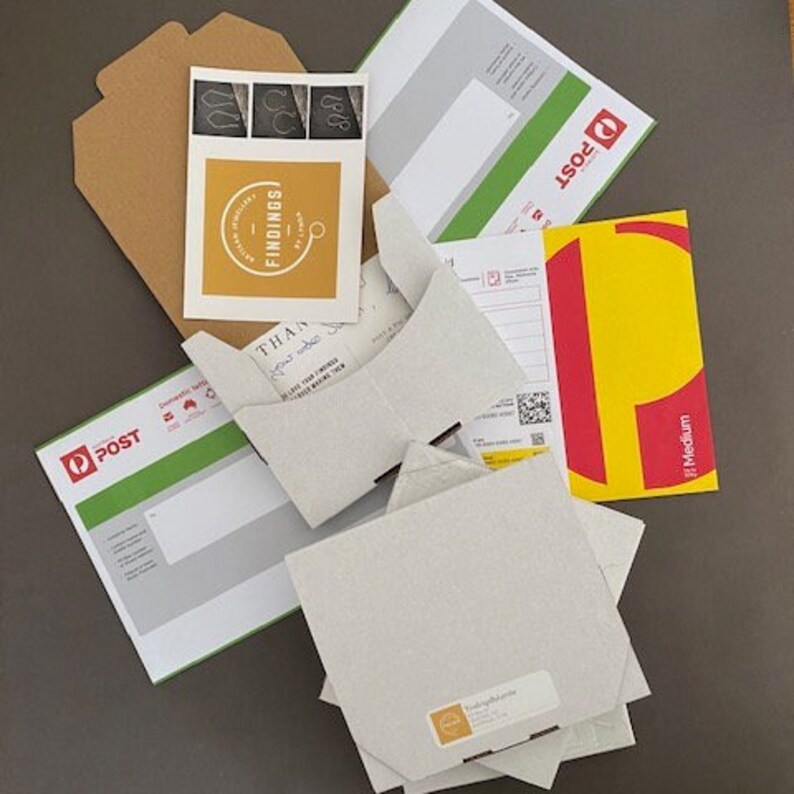 May include: A flat cardboard box with a white label that says "Thank you" and "Please leave your printed address details below". The box is surrounded by various packaging materials, including a brown envelope, a green and white envelope with the word "POST" on it, and a yellow and red envelope with the word "POST" on it.