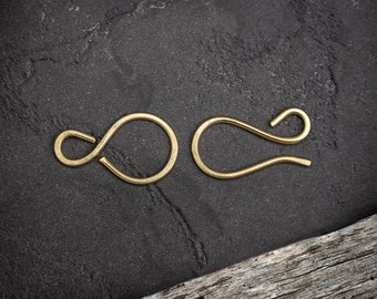 May include: Two gold-colored, handmade jewelry clasps. One clasp is a circle with a loop, and the other is a curved hook. The clasps are displayed on a dark, textured surface with a piece of driftwood in the corner.