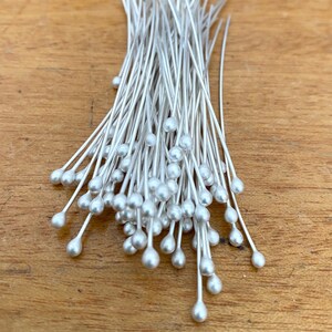 May include: A bunch of silver-toned, teardrop-shaped head pins with long, thin stems. The pins are arranged in a loose pile.