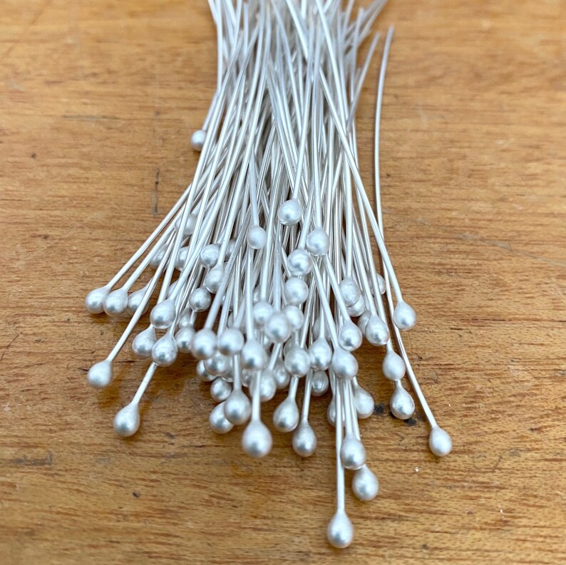 May include: A bunch of silver-toned, teardrop-shaped head pins with long, thin stems. The pins are arranged in a loose pile.