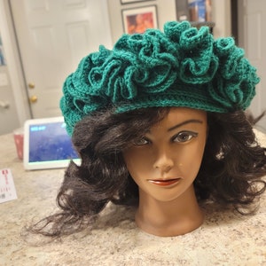May include: A handmade, emerald green crochet hat with a ruffled design. The hat is displayed on a mannequin head with dark brown hair. The hat's texture and color are prominent, showcasing the craftsmanship of the item.