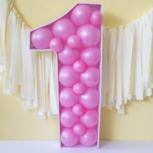 May include: A pink number one balloon decoration made of many smaller pink balloons. The number one is standing on a white surface in front of a yellow wall.