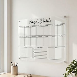 Peut inclure: Un calendrier mural en acrylique transparent avec l'inscription "Kenzie's Schedule" en écriture cursive. Il comporte des sections pour chaque jour de la semaine, des rappels, un planificateur de repas et des notes. Il est fixé sur un mur blanc.
