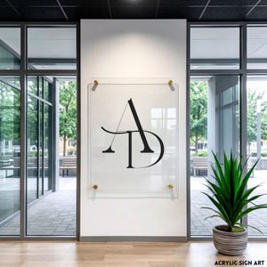 Könnte beinhalten: Klarer Acrylschild mit den schwarzen Buchstaben "AD", montiert an einer weißen Wand mit goldfarbenen Beschlägen. Das Schild befindet sich in einer modernen Büroumgebung mit großen Fenstern und einer Topfpflanze.