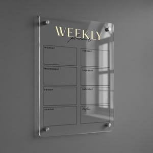 Puede incluir: Un planificador semanal de acrílico transparente con letras doradas y un diseño minimalista. El planificador está dividido en secciones para cada día de la semana, con espacio para notas. Está montado en una pared gris.