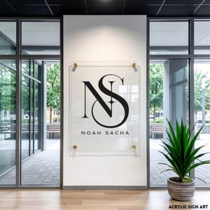 May include: Clear acrylic sign featuring the black initials "NS" and the name "NOAH SACHA" in a contemporary font. The sign is mounted on a white wall with gold-coloured fixings. The sign is in an office setting with large windows and a potted plant.