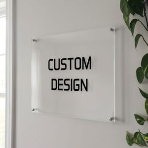 May include: A clear acrylic sign with the words "CUSTOM DESIGN" in bold, black letters. The sign is mounted on a white wall with silver hardware. Green plants are visible in the background.