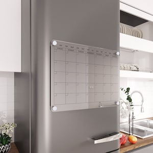Puede incluir: Un calendario de borrado en seco de acrílico transparente con un marco blanco y letras negras. El calendario está montado en un refrigerador de acero inoxidable con imanes. El calendario tiene los meses del año listados en la parte superior y los días de la semana listados en el lateral.
