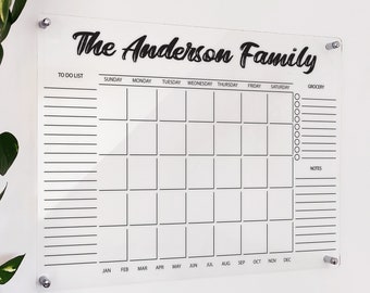 May include: A clear acrylic dry-erase calendar with a black border and black text. The calendar is labelled with the days of the week and months of the year. The top of the calendar says "The Anderson Family".