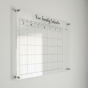 May include: A clear acrylic family calendar with the words "Our Family Calendar" in black script. The calendar grid is divided into weeks and months, with the days of the week labeled across the top. Silver hardware secures the calendar to the wall.
