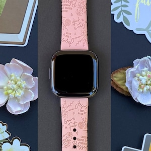 May include: A pink Apple Watch band with a floral design and inspirational quotes, including "Jesus Loves You", "Grow in Faith", "Created with a Purpose", and "Pray More Worry Less".
