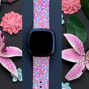 Puede incluir: Una correa de reloj con estampado floral rosa y azul con una esfera negra. La correa est&aacute; hecha de silicona y tiene un dise&ntilde;o floral con lirios rosas sobre un fondo azul claro.