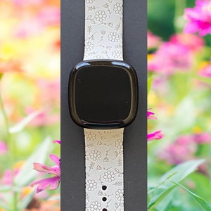 K&ouml;nnte beinhalten: Wei&szlig;es Silikon-Armband mit Blumenmuster. Das Armband ist an einer schwarzen Smartwatch mit rundem Zifferblatt befestigt.