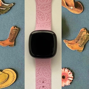 May include: A pink smartwatch band with a paisley pattern. The band is shown on a white background with various decorative items, including pink flowers, cowboy hats, and cowboy boots.