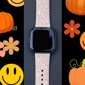 Pu&ograve; includere: Un cinturino rosa per smartwatch con sfondo bianco e un motivo di facce sorridenti e zucche. Il cinturino &egrave; su uno sfondo nero.