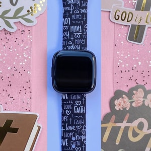 K&ouml;nnte beinhalten: Schwarz-wei&szlig;es Apple Watch Armband mit einem sich wiederholenden Muster der W&ouml;rter "Glaube", "Liebe" und "Hoffnung". Das Armband befindet sich auf einem rosa Hintergrund mit anderen religi&ouml;sen Aufklebern.