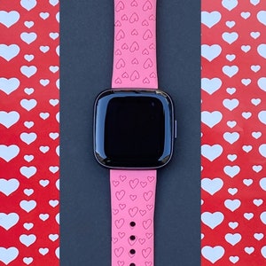 May include: A pink silicone watch strap with a heart pattern. The strap is designed for a smartwatch and has a black screen.