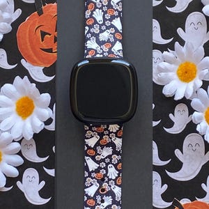 May include: Black smartwatch with a Halloween-themed band featuring white ghosts, orange pumpkins, and small flowers on a dark background. The band is made of a flexible material and is designed to fit a smartwatch. The background includes white daisies and ghost cutouts.