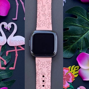 May include: A pink Apple Watch band with a flamingo and floral pattern. The band is shown on a black background with pink flowers and a pineapple.