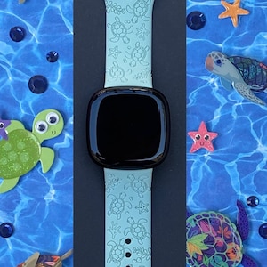 May include: A light blue silicone watch band with a pattern of sea turtles. The band is designed for a smartwatch and has a black watch face.